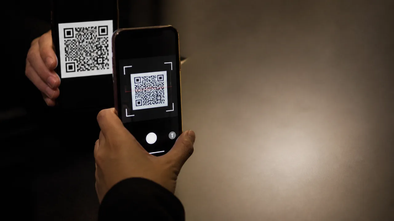 A glowing arrow points from a smartphone displaying a QR code to a computer screen showing binary data, set against a dark…