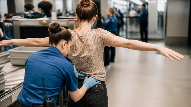 Four travelers with red X marks on their outfits stand in an airport terminal while a woman gestures disapprovingly beside…